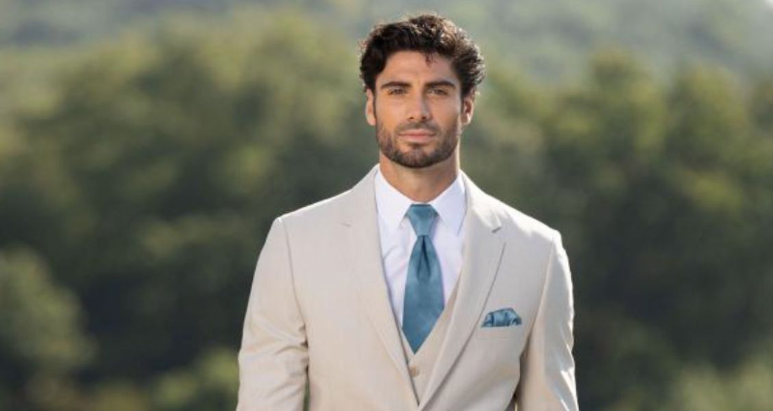 Groom wearing a tan summer wedding tuxedo with a teal tie and matching pocket square, standing outdoors with trees in the background.
