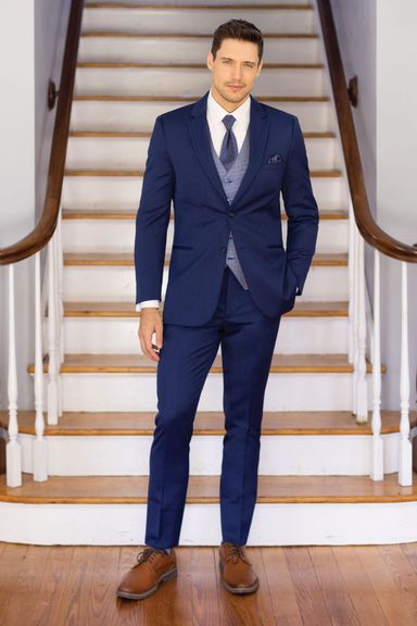 Groom in a navy blue summer tuxedo with a light blue vest and dark tie, standing confidently on an indoor staircase.