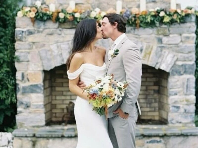 Close-up of a bride and groom kissing in front of a stone fireplace; the bride wears a minimalist off-the-shoulder gown with timeless charm.