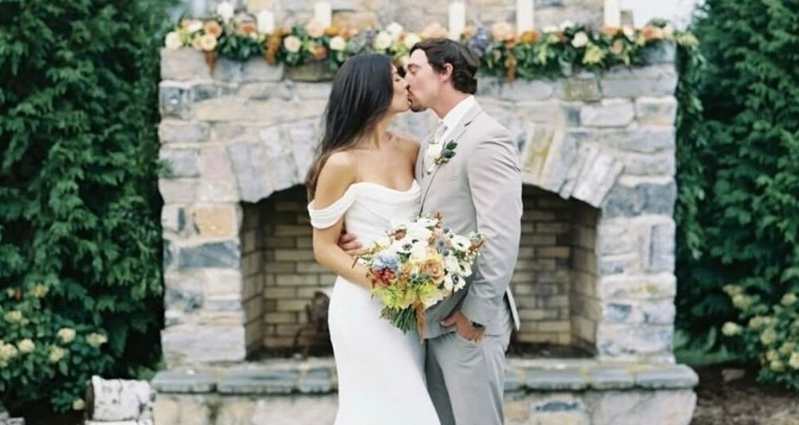 Close-up of a bride and groom kissing in front of a stone fireplace; the bride wears a minimalist off-the-shoulder gown with timeless charm.
