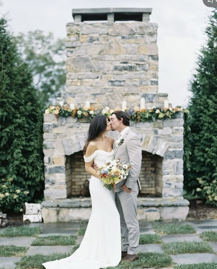 Bride and groom share a kiss in front of a stone fireplace; the bride wears an off-the-shoulder fitted gown, evoking timeless elegance.