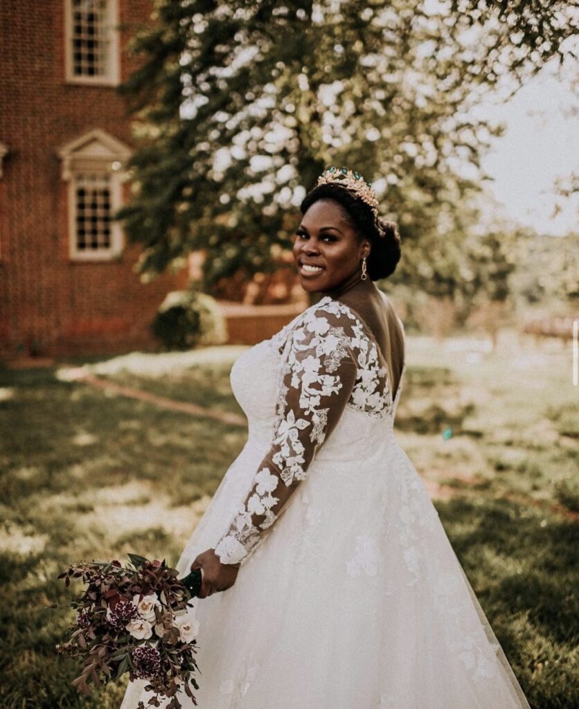 Bride smiles over her shoulder outdoors in a lace-sleeved ballgown wedding dress with floral details and a regal crown, holding a moody bouquet.