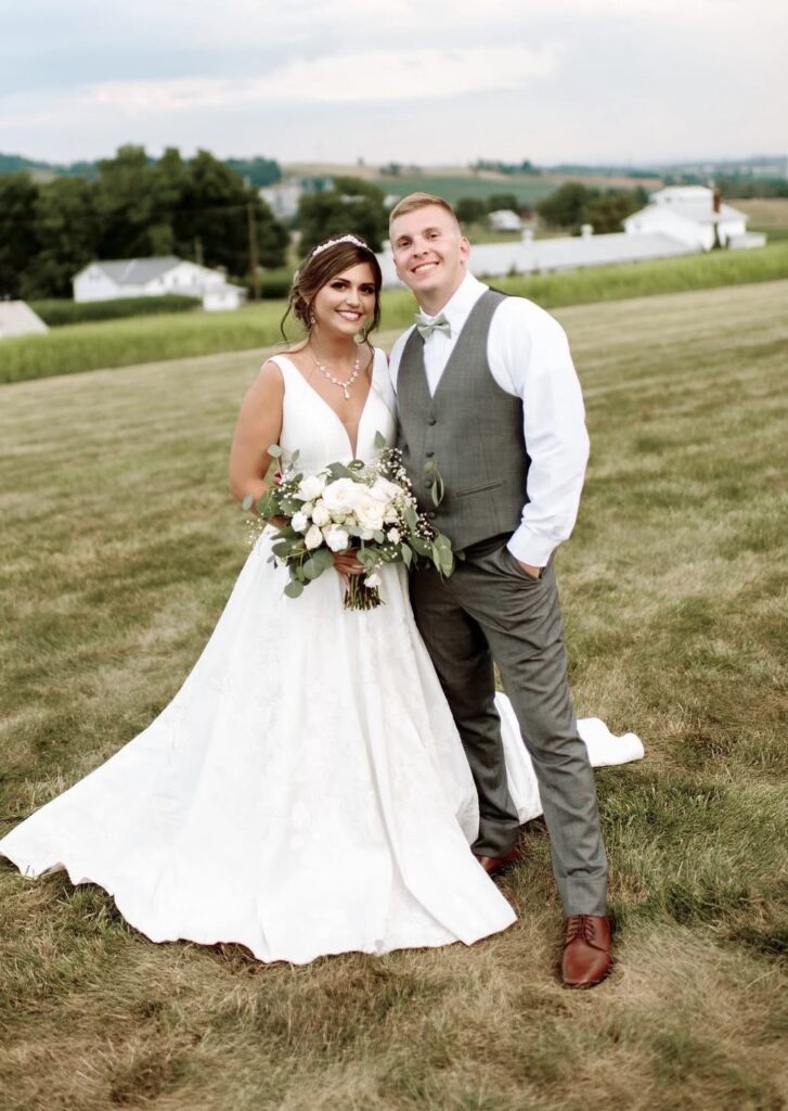 Bride and groom pose together in an open field; the bride wears a sleeveless A-line gown with a plunging neckline and delicate accessories.