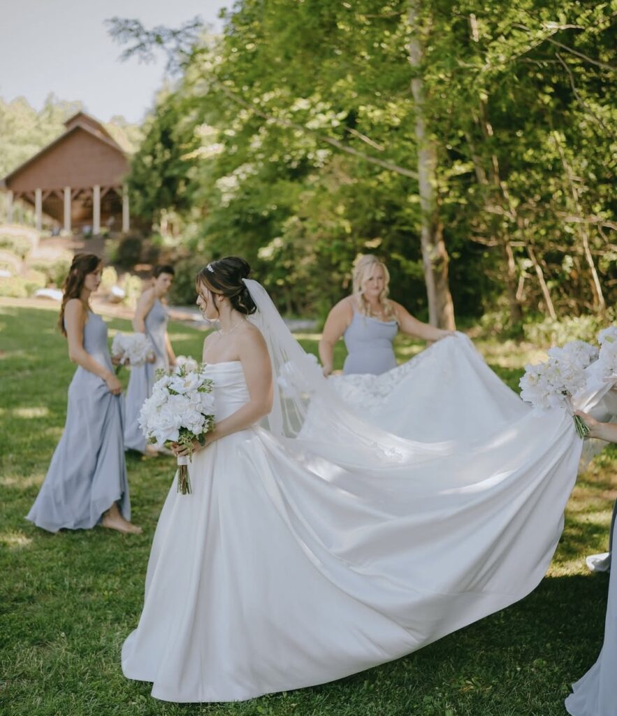 Bride walks through lush greenery with bridesmaids helping carry her long train; she wears a strapless satin ballgown with a long veil.