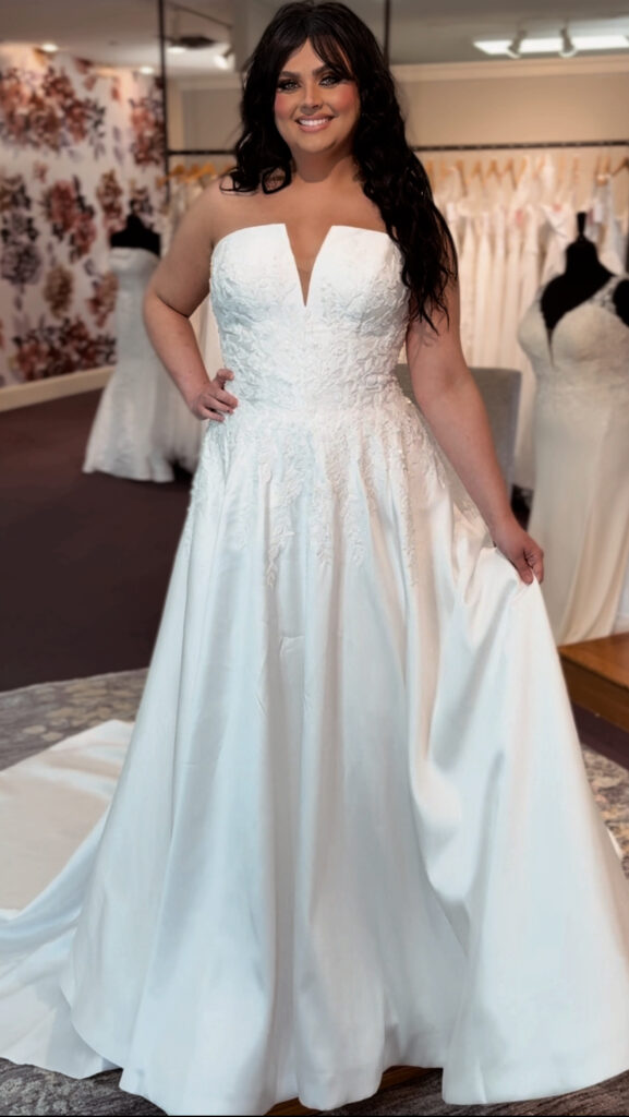 Full view of a bride in a strapless ballgown wedding dress with a deep neckline and embroidered bodice, posing inside Amanda’s Touch bridal boutique.