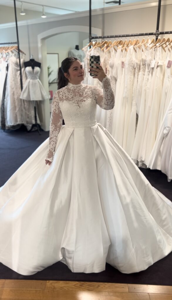 Bride taking a mirror selfie in a dramatic ballgown wedding dress with a high lace neckline and long lace sleeves in a bridal showroom.