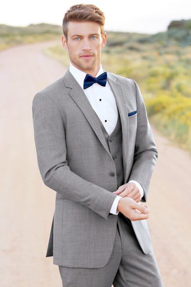 Groom modeling a heather grey wedding tuxedo with a navy bowtie and white shirt, walking on a rural dirt path.