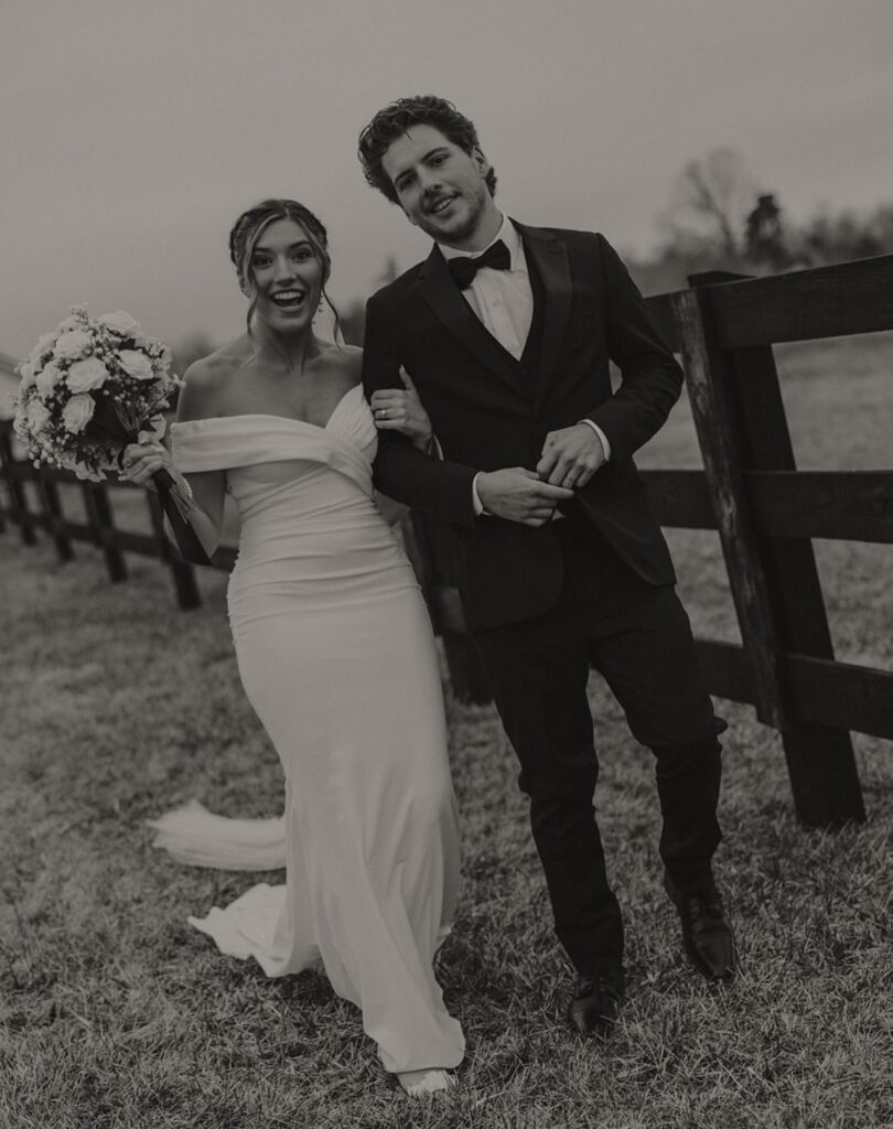 A black-and-white photo of a bride and groom walking along a wooden fence, with the bride in an off-the-shoulder dress holding a bouquet.