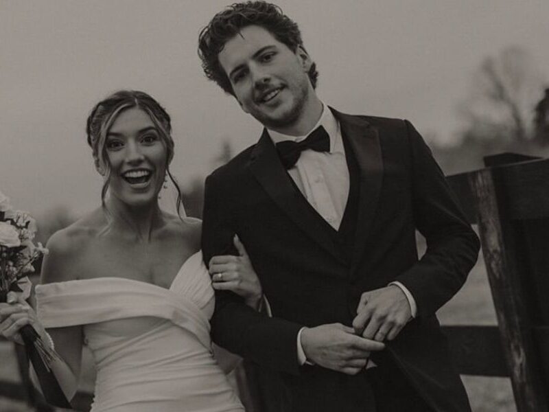 A black-and-white photo of a bride and groom walking along a wooden fence, with the bride in an off-the-shoulder dress holding a bouquet.