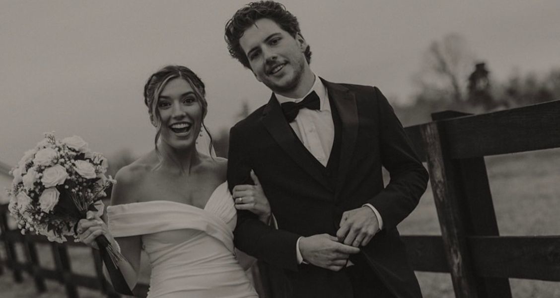 A black-and-white photo of a bride and groom walking along a wooden fence, with the bride in an off-the-shoulder dress holding a bouquet.