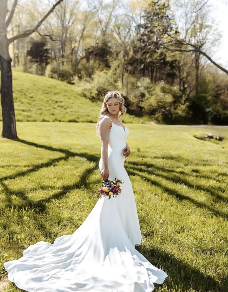 A bride stands on a sunny lawn holding a bouquet of colorful flowers, wearing a sleek, simple gown with a long train and fitted bodice.