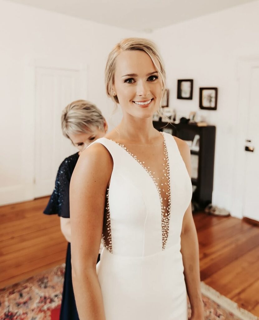 A smiling bride with blonde hair in an updo wears a minimalist wedding gown with a deep V-neckline accented by beaded detailing.