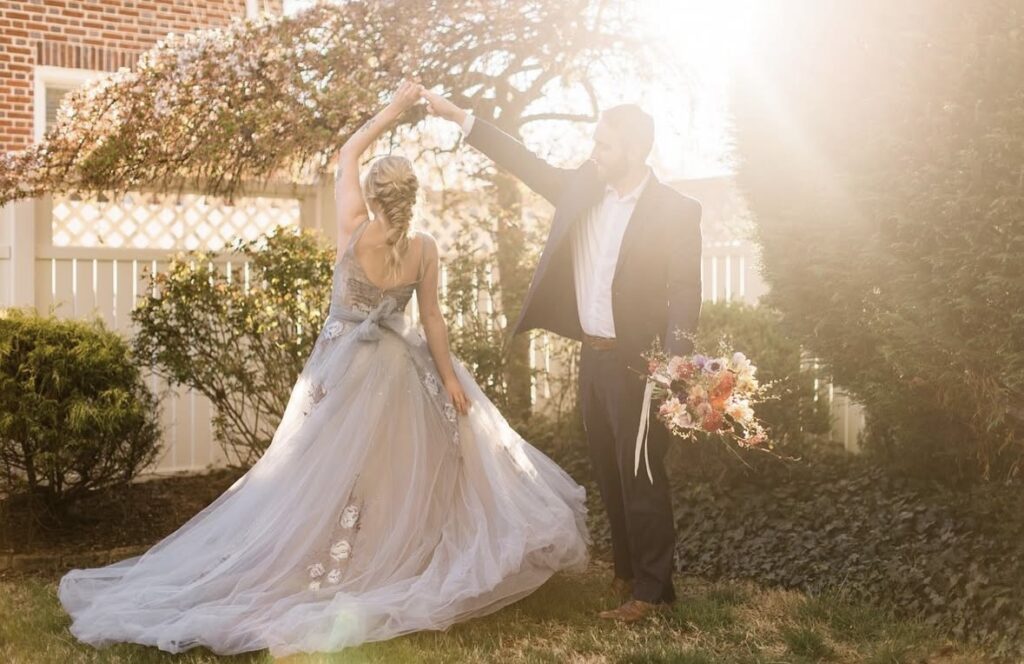 A bride in a gray-blue floral applique wedding gown twirls with her groom in a sunlit backyard garden as he holds a bouquet of colorful spring flowers.