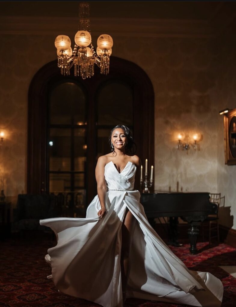 A bride twirls joyfully in an elegant strapless gown with a slit, in a candlelit ballroom beneath chandeliers and a grand piano.
