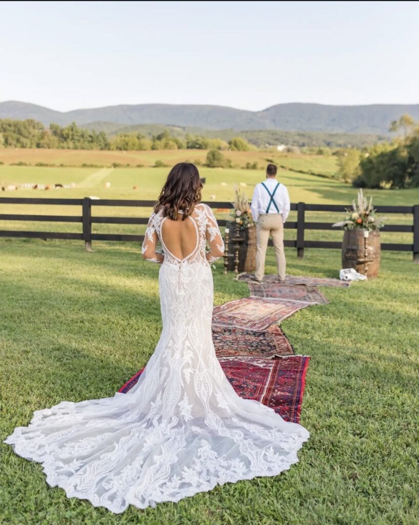 A bride wearing a lace mermaid gown with an open back walks toward her groom, who stands on vintage rugs in front of a scenic Virginia countryside backdrop.