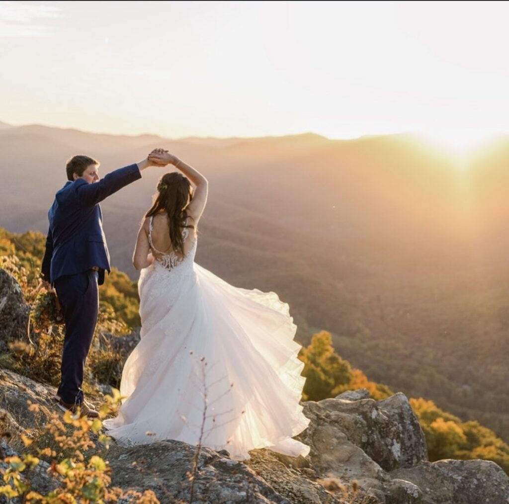 A bride in a flowing tulle ballgown dances with her groom on a mountaintop at sunset, overlooking rolling hills in the distance.