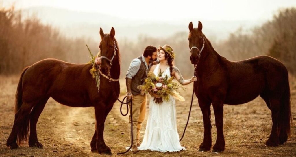 A couple poses between two brown horses on a country trail, with the bride holding a boho bouquet and wearing a lace gown and sunhat.