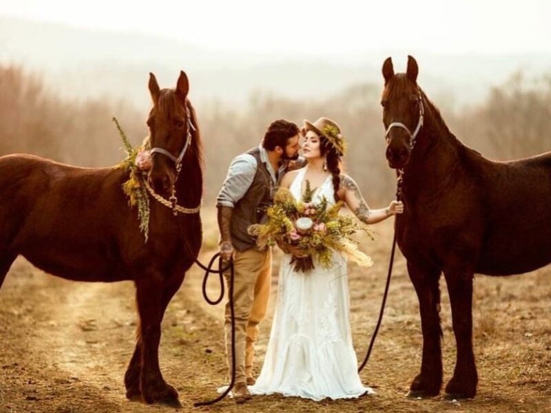 A couple poses between two brown horses on a country trail, with the bride holding a boho bouquet and wearing a lace gown and sunhat.