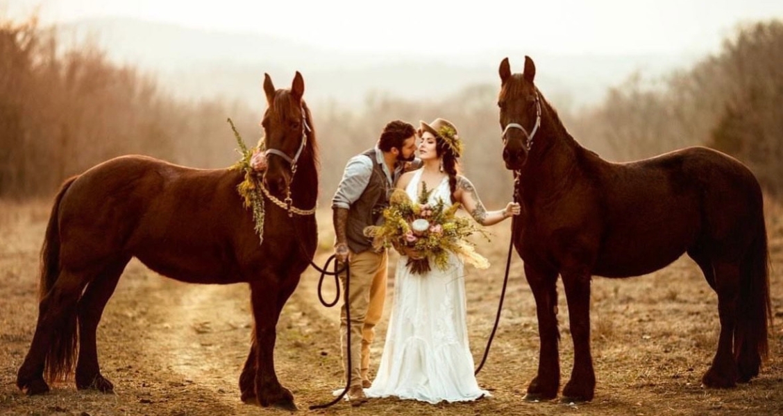 A couple poses between two brown horses on a country trail, with the bride holding a boho bouquet and wearing a lace gown and sunhat.