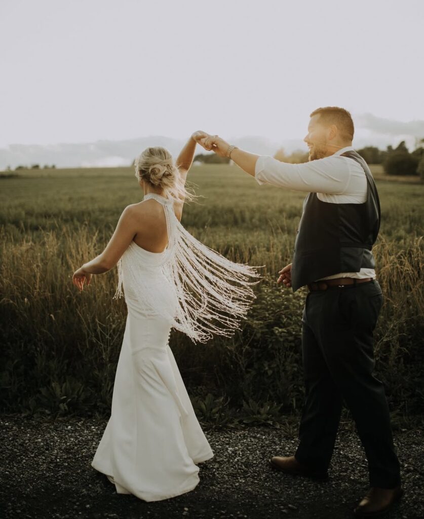 A bride dances with her groom in the countryside at sunset, wearing a sleek white wedding gown with vintage-inspired fringe detailing.