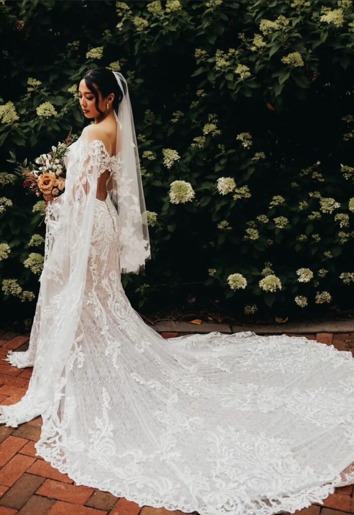 A bride poses in front of lush hydrangeas, wearing a fitted lace wedding gown with dramatic sleeves and a cathedral-length train.