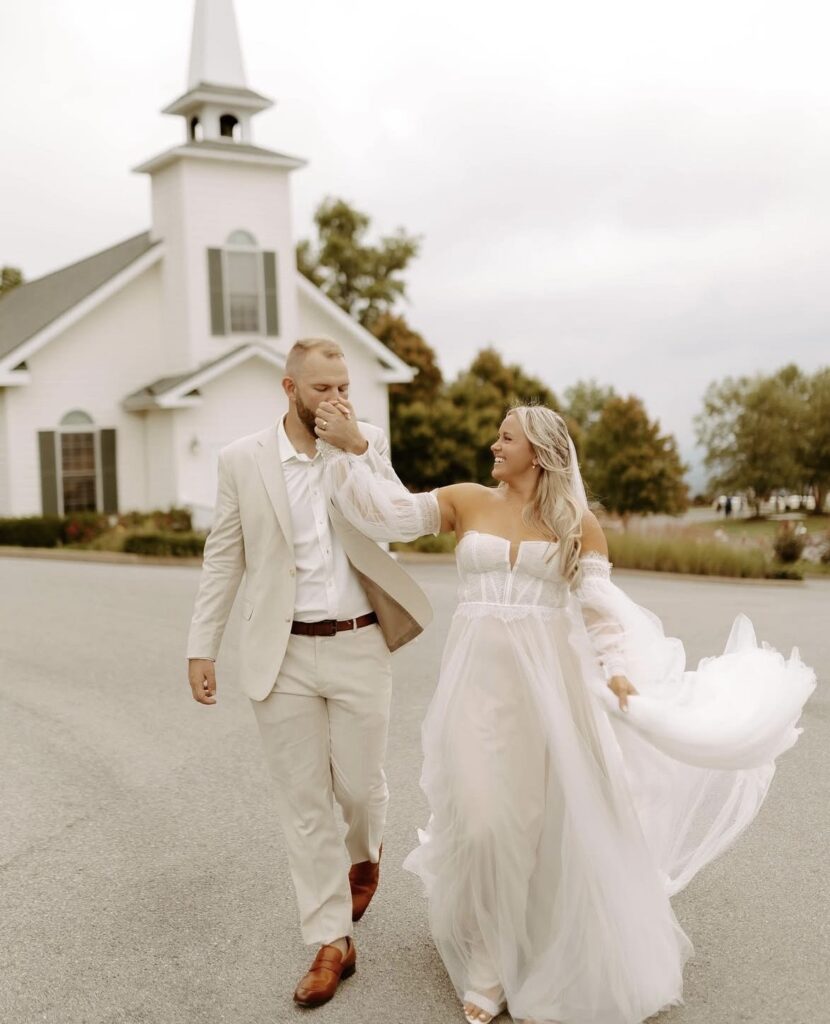 A smiling couple walks hand in hand outside a chapel, with the bride in a sheer tulle dress with off-the-shoulder sleeves and corset bodice.
