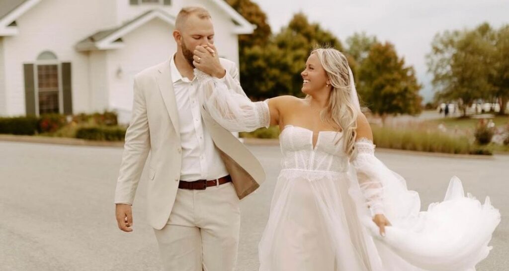 A groom gently kisses the bride’s hand as she walks in a flowing, vintage-inspired gown with sheer sleeves and a romantic silhouette.