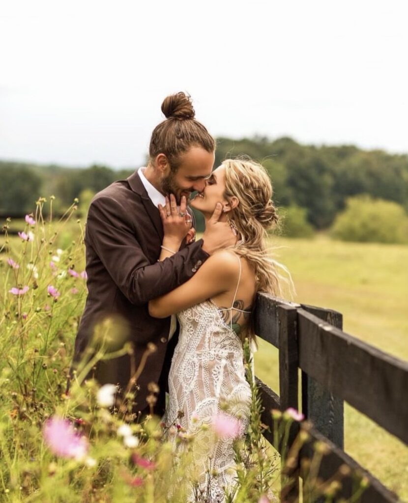 A bride stands beside a groom in a wildflower field, wearing a vintage lace gown with thin straps and intricate detailing as they kiss.