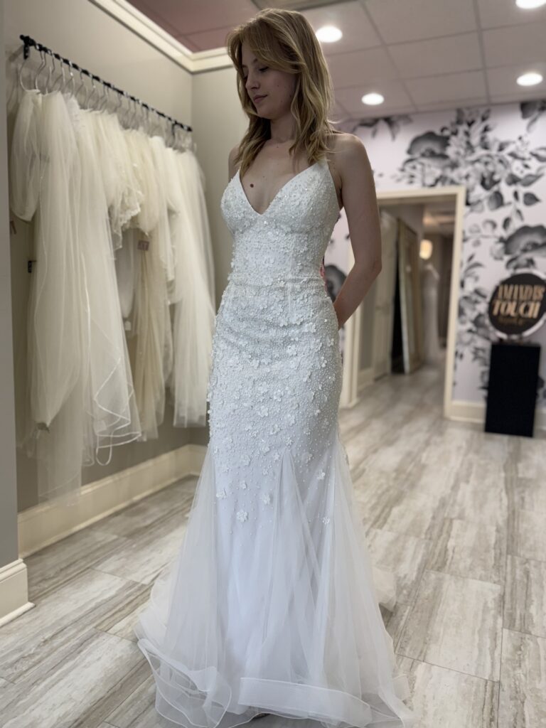 A bride in a sleeveless V-neck wedding gown with floral appliqué and a tulle skirt, standing beside veils in the Harrisonburg showroom.