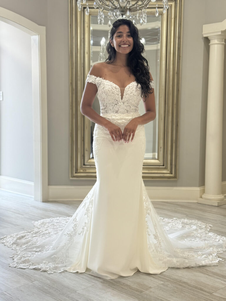 A bride in a fitted off-the-shoulder lace gown with a deep neckline and floral details, standing beneath a chandelier at Amanda's Touch.