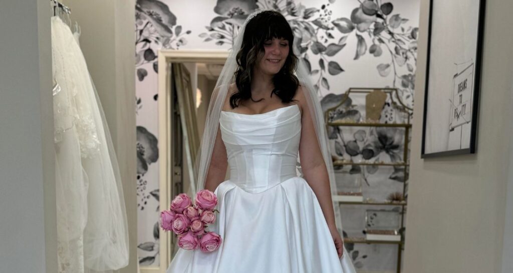 A bride smiling while holding a bouquet of pink roses, wearing a basque waist wedding gown with a folded pleated bodice and long train.