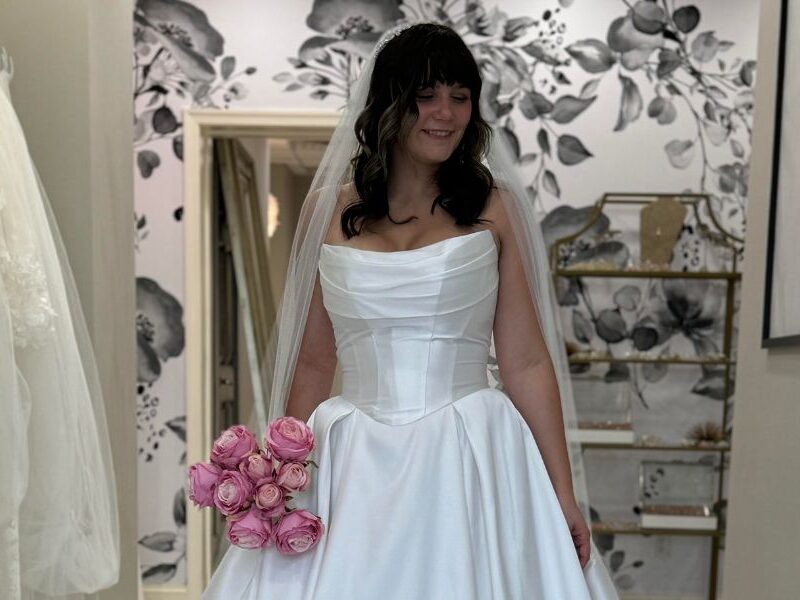 A bride smiling while holding a bouquet of pink roses, wearing a basque waist wedding gown with a folded pleated bodice and long train.