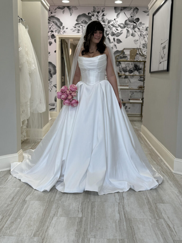 A bride wearing a structured satin basque waist wedding dress with a pleated neckline, holding pink roses and smiling in a boutique.