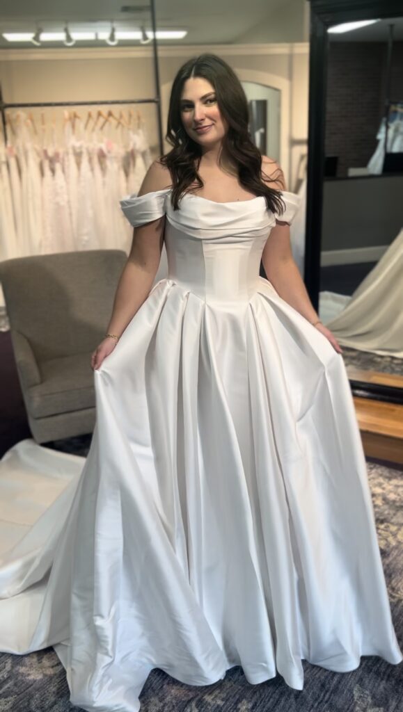 A bride in an off the shoulder satin ball gown wedding dress with a defined basque waist, standing in a softly lit bridal showroom.