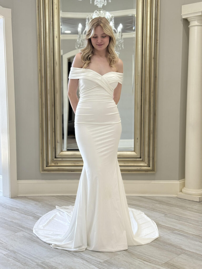 A bride in a sleek, off-the-shoulder fitted gown with a slight flare at the hem, standing in front of a gold mirror and chandelier display.