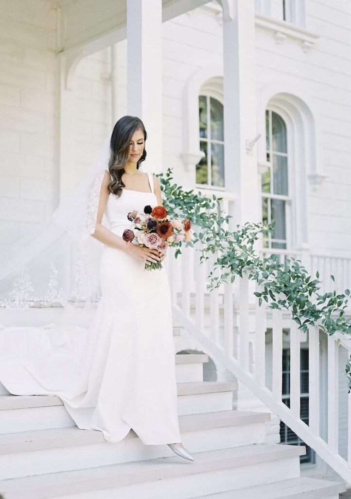A bride in a minimalist square-neck wedding dress descends white mansion steps, holding a bouquet of red, pink, and plum flowers.
