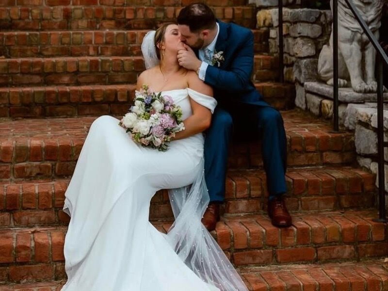A couple sits on brick steps outside a historic mansion, the bride in an off-the-shoulder wedding gown and the groom kissing her cheek.