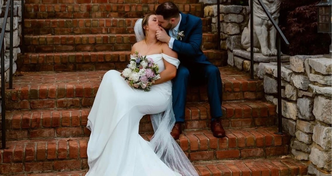 A couple sits on brick steps outside a historic mansion, the bride in an off-the-shoulder wedding gown and the groom kissing her cheek.