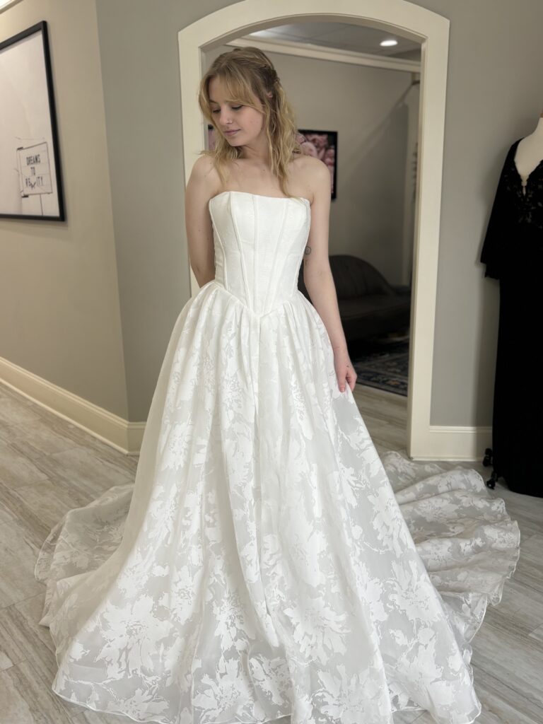 A bride in a strapless floral jacquard wedding dress with a basque waist and flowing train, posing near a full-length mirror.