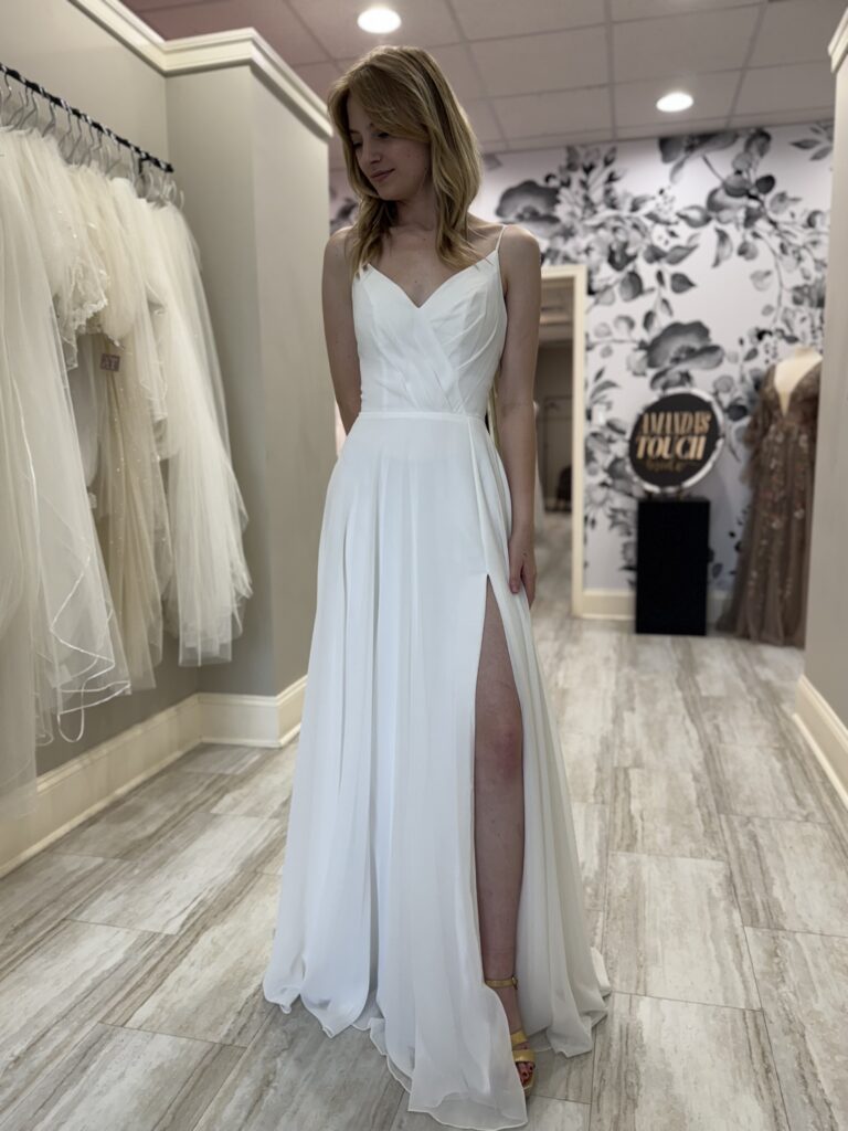 A bride in a lightweight chiffon wedding dress with spaghetti straps and a front leg slit, standing near veils at Amanda's Touch in Harrisonburg.