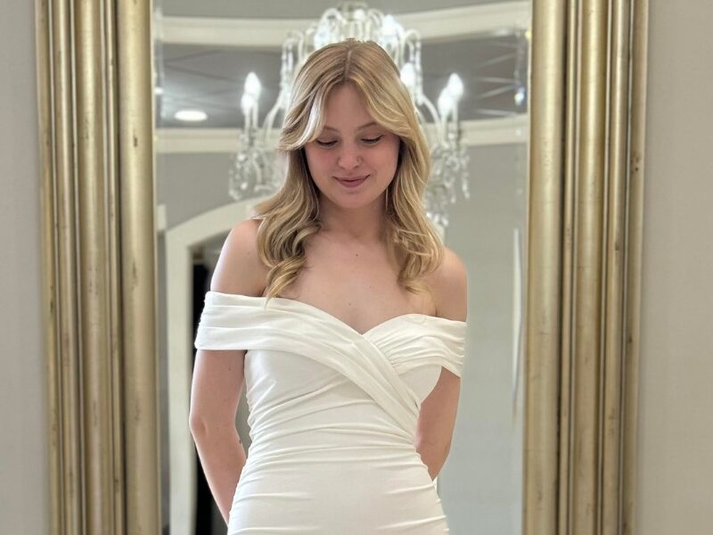 A bride wearing an off-the-shoulder fitted wedding dress with a ruched bodice and a soft train, standing in front of a gold-framed mirror.