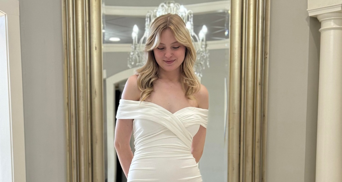 A bride wearing an off-the-shoulder fitted wedding dress with a ruched bodice and a soft train, standing in front of a gold-framed mirror.