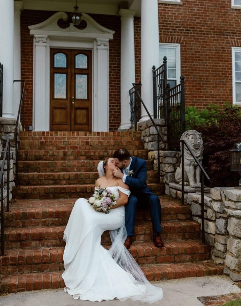 A bride and groom share a kiss on brick steps in front of a mansion with white columns and double wooden doors, framed by lion statues.