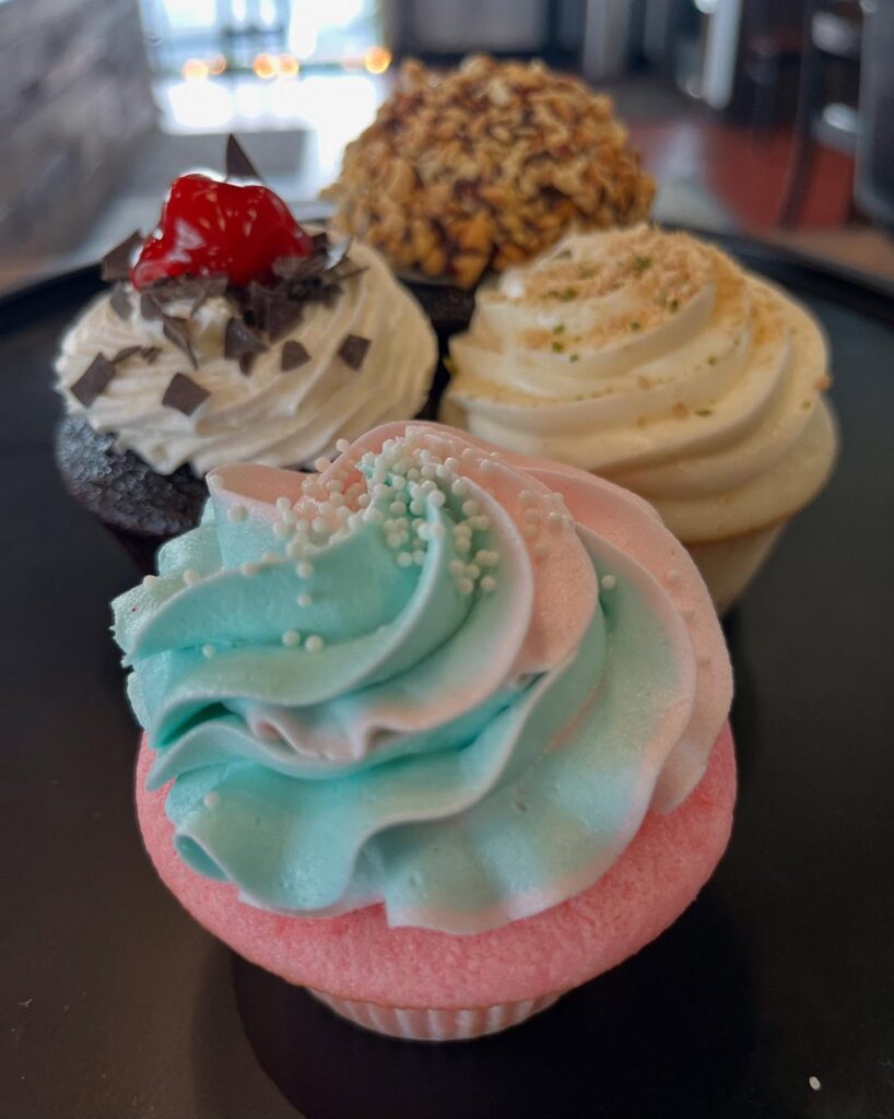 A variety of colorful cupcakes are arranged on a tray, including a pink and blue swirl cupcake, one with a cherry and chocolate shavings, and another with chopped nuts.