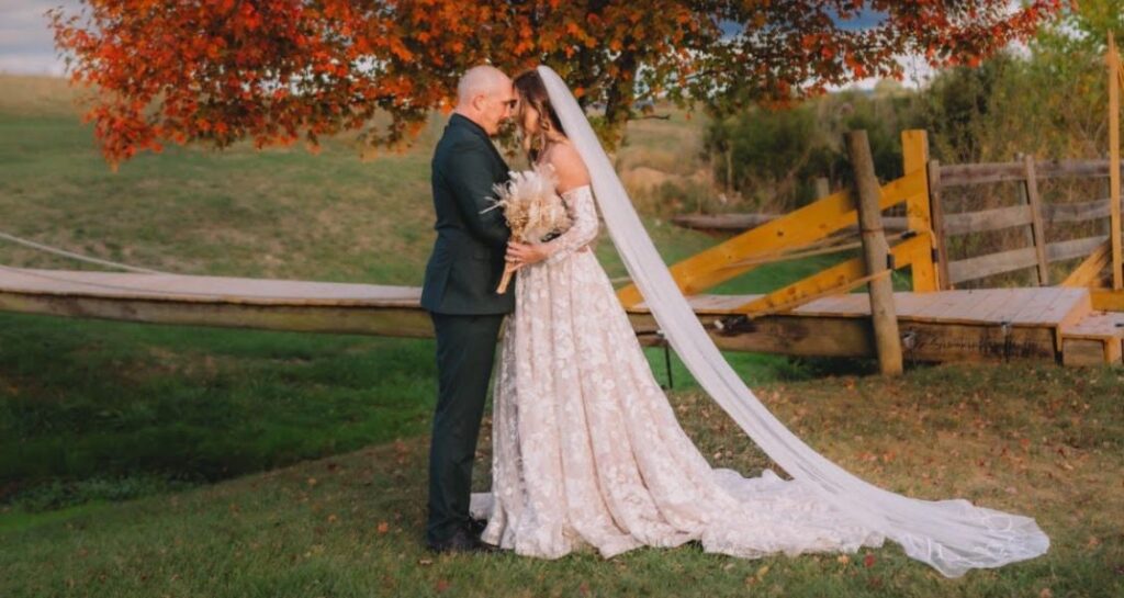 A bride in an off-the-shoulder floral gown stands with her groom under a vibrant red-orange tree in a countryside setting.