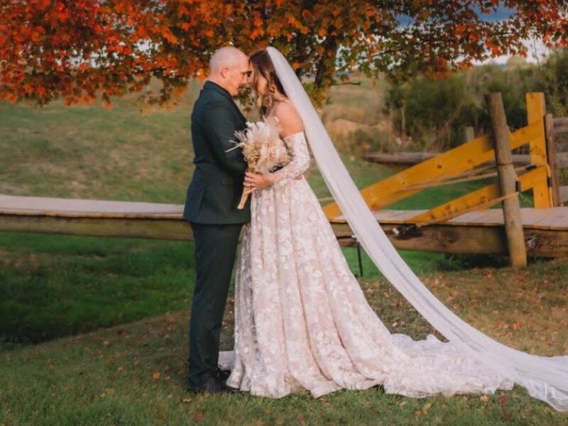 A bride in an off-the-shoulder floral gown stands with her groom under a vibrant red-orange tree in a countryside setting.