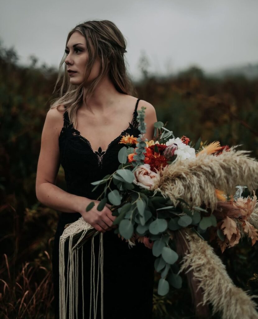 A close-up of a bride holding a bouquet with pampas grass and fall flowers, wearing a black lace gown in a moody outdoor setting.