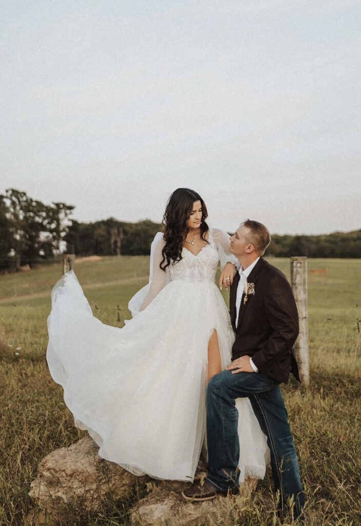 A bride in a long sleeve white gown with a thigh-high slit stands beside her groom in a country field at sunset.