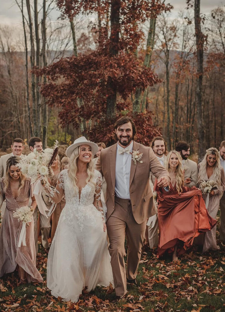 A joyful bride and groom walk with their wedding party through autumn leaves, with the bride in a long sleeve lace dress and the groom in a tan suit.