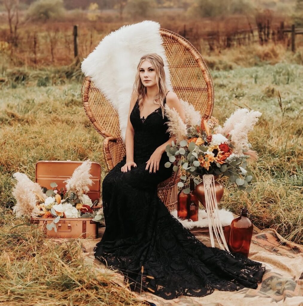 A bride in a black lace wedding gown sits in a wicker chair surrounded by pampas grass and fall florals in a rustic outdoor field.